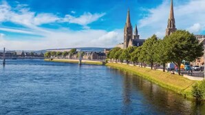 Ein Fluss mit einer Brücke, eine Stadtansicht mit zwei markanten Türmen und eine grüne Parkanlage mit Bäumen.