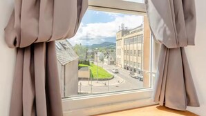 Ein Zimmer mit großem Fenster, Vorhängen und Blick auf eine Straße mit Gebäuden und Grünflächen.