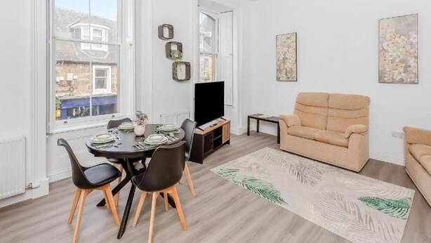 Un salon moderne avec un canapé beige, une table à manger dressée pour quatre personnes et une grande fenêtre donnant sur les bâtiments environnants.