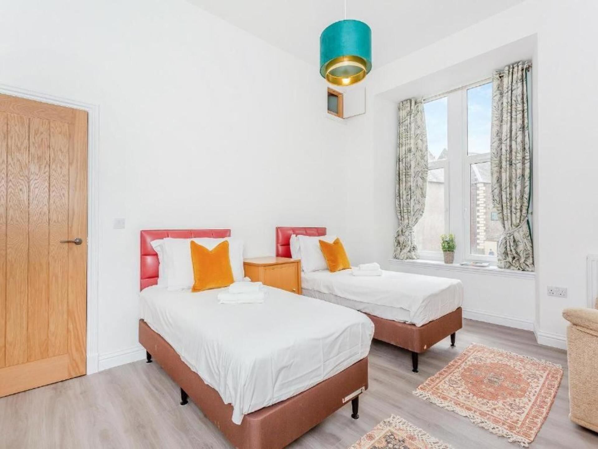A bedroom with two beds, a wooden door, a window with curtains, and a hanging light fixture.