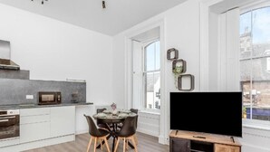 Une cuisine moderne avec des meubles blancs, une table à manger avec des chaises, un téléviseur à écran plat posé sur un meuble en bois et de grandes fenêtres.