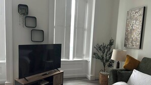 Un salon moderne avec un téléviseur à écran plat posé sur un meuble en bois, un canapé avec des coussins, une table d’appoint avec une lampe et une plante décorative.