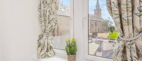 A window with a view of a church tower, curtains, a small plant, and a magazine on the windowsill.