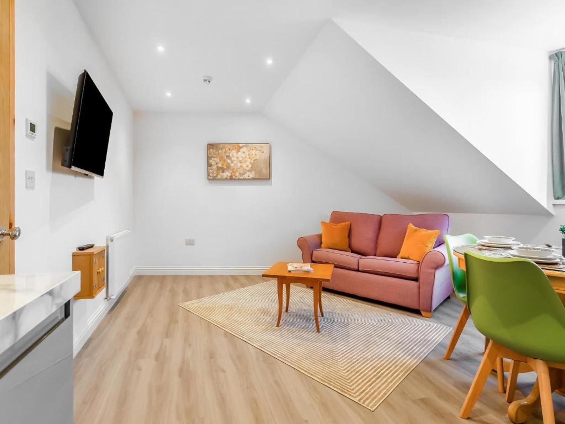 Ein modernes Wohnzimmer mit schrägverdeckter Decke, einem rosa Sofa mit orangenen Kissen, einem Holztisch und einem grünen Sessel.