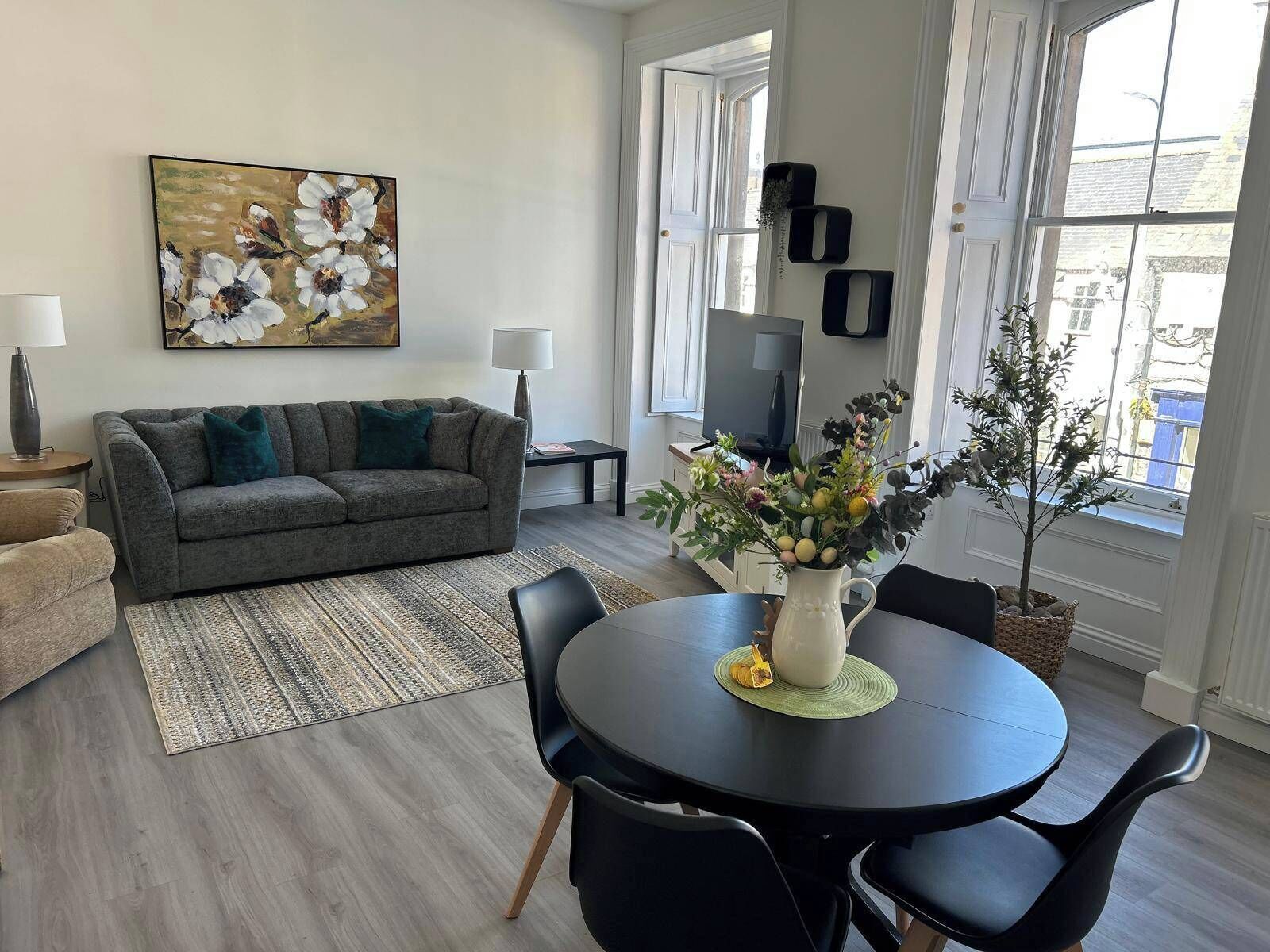 A modern living room with a grey sofa, a round dining table with black chairs, a coffee table with a vase of flowers, and a painting on the wall.