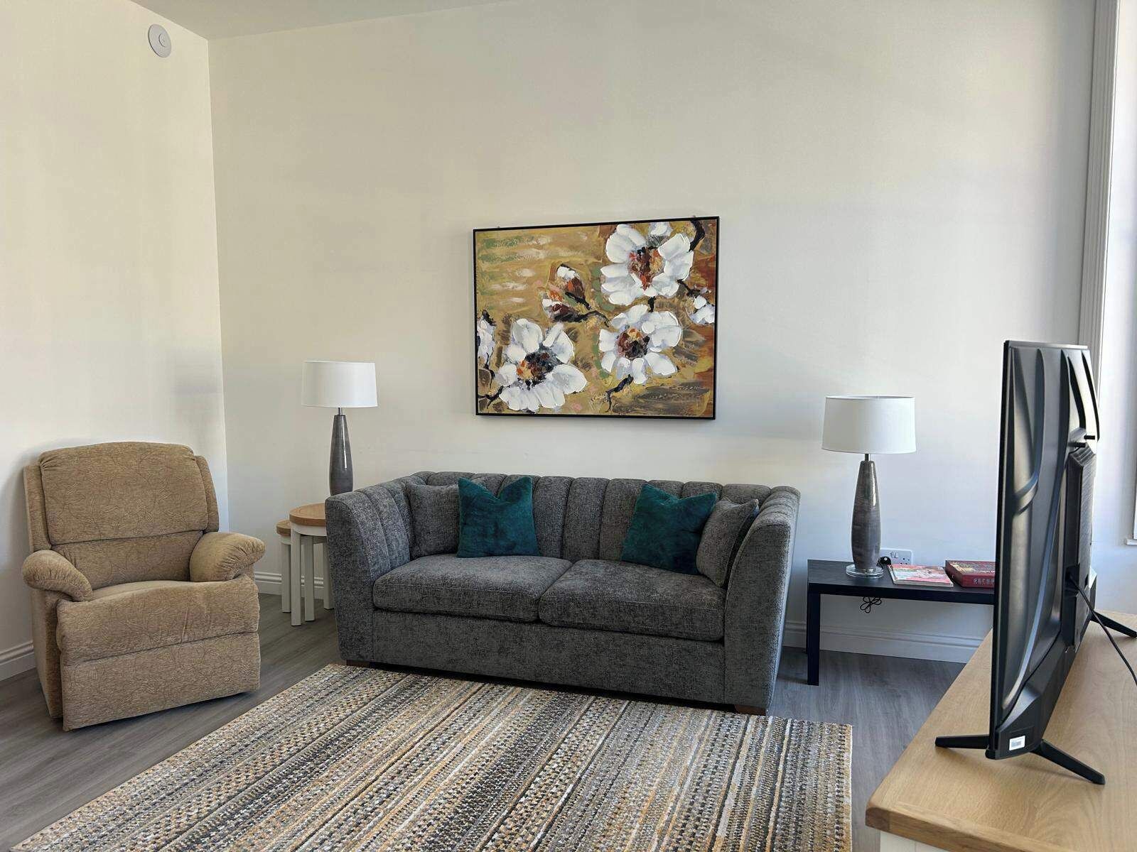 A living room with a grey sofa, a beige armchair, a patterned rug, a painting of flowers, a lamp, and a TV on a wooden stand.
