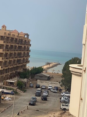 A view of a coastal area with a parking lot, a beach, and a multi-story building with balconies. - Spacious 3-bedroom apartment in soothing Hurghada with AC (Hurghada)