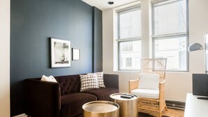 Un salon moderne avec un mur bleu foncé, un canapé marron, un fauteuil blanc et deux petites tables d’appoint à bordure dorée.