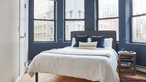 Une chambre à coucher avec un grand lit, des murs bleu foncé et de nombreuses fenêtres donnant sur la ville.