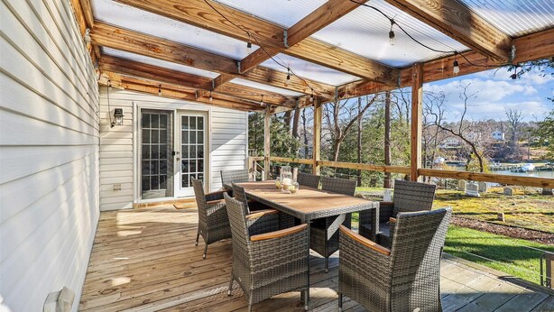 En overdækket terrasse med et træbord og fletstole, med udsigt over en græsplæne med træer og et hus i det fjerne.