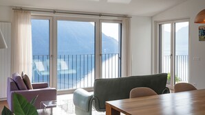 Un soggiorno con divano, poltrone, un tavolo in legno e grandi porte scorrevoli che si aprono su un balcone con vista sul mare e sulle montagne.