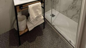 Een moderne badkamer met een glazen douchecabine, een betegelde wand van marmer, een witte wastafel en een spiegel erboven.