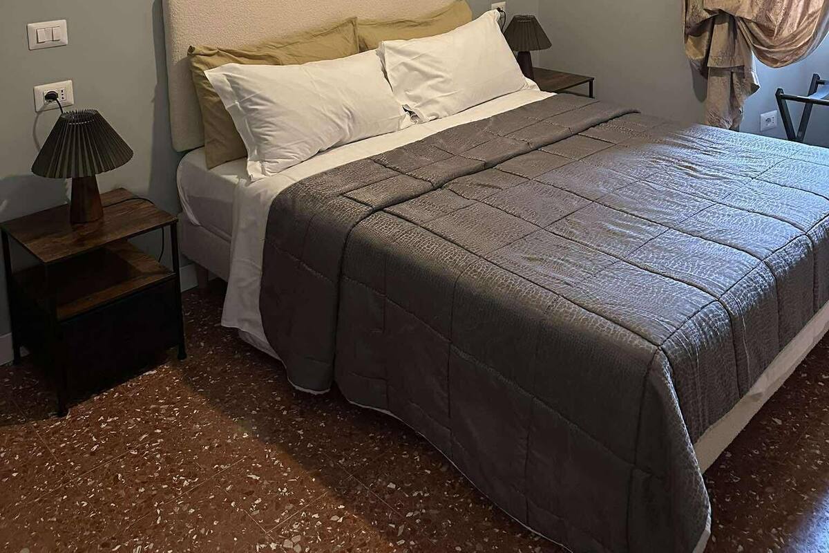Ein Schlafzimmer mit Bett, Nachttisch, Lampe und Vorhang.