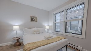 A neatly arranged bedroom with a bed, two bedside lamps, a window with blinds, and a small plant on the windowsill. - Theater District Cozy 1 BR! (Boston)