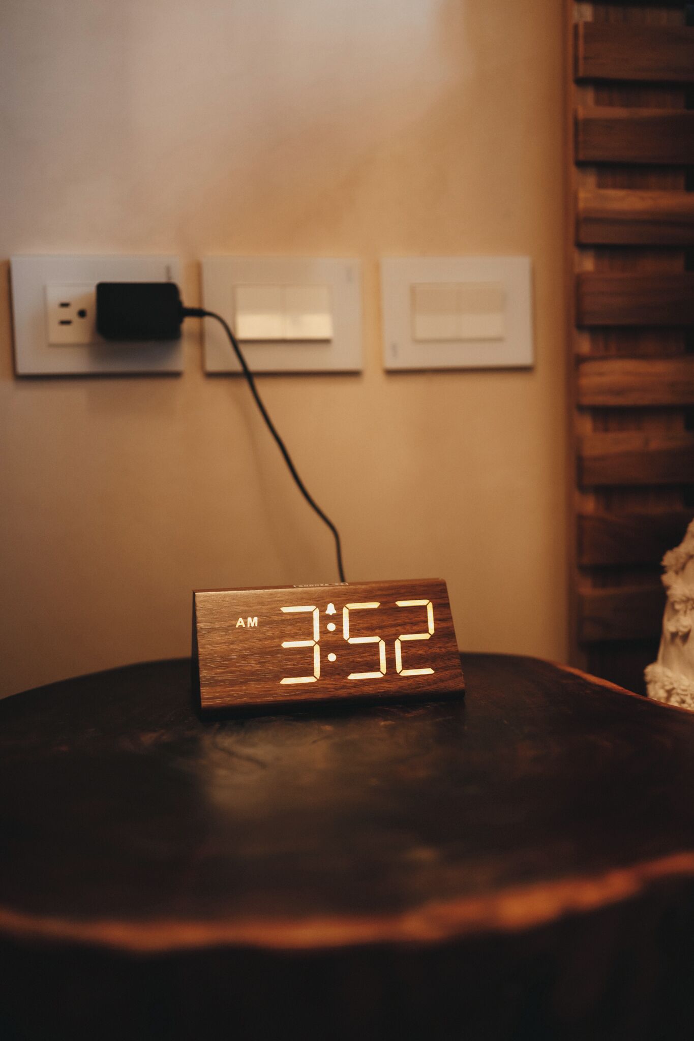 Un reloj digital de madera muestra las 3:52 de la madrugada sobre una mesa, con un enchufe de corriente al fondo.
