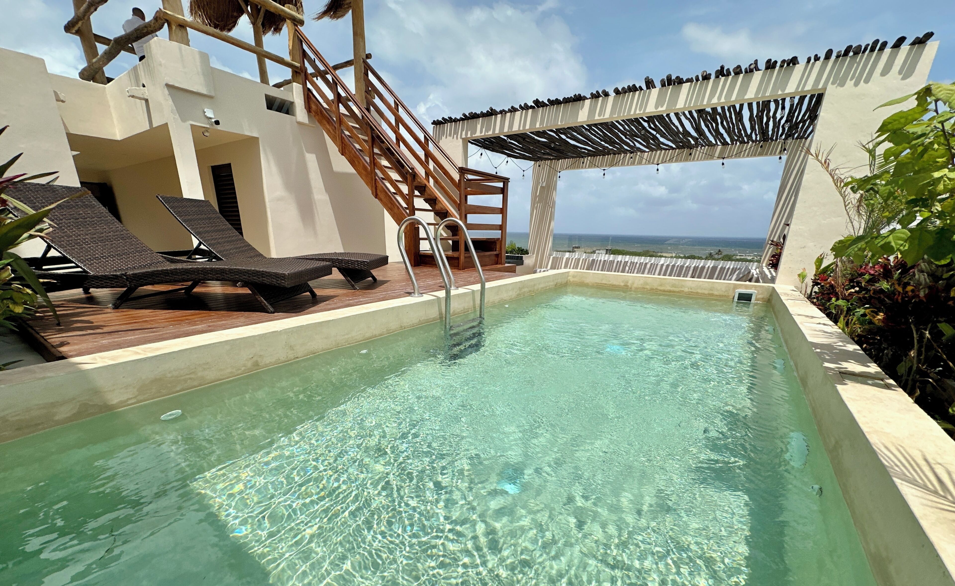 A clear pool with wooden loungers, a wooden staircase, and a view of the ocean.