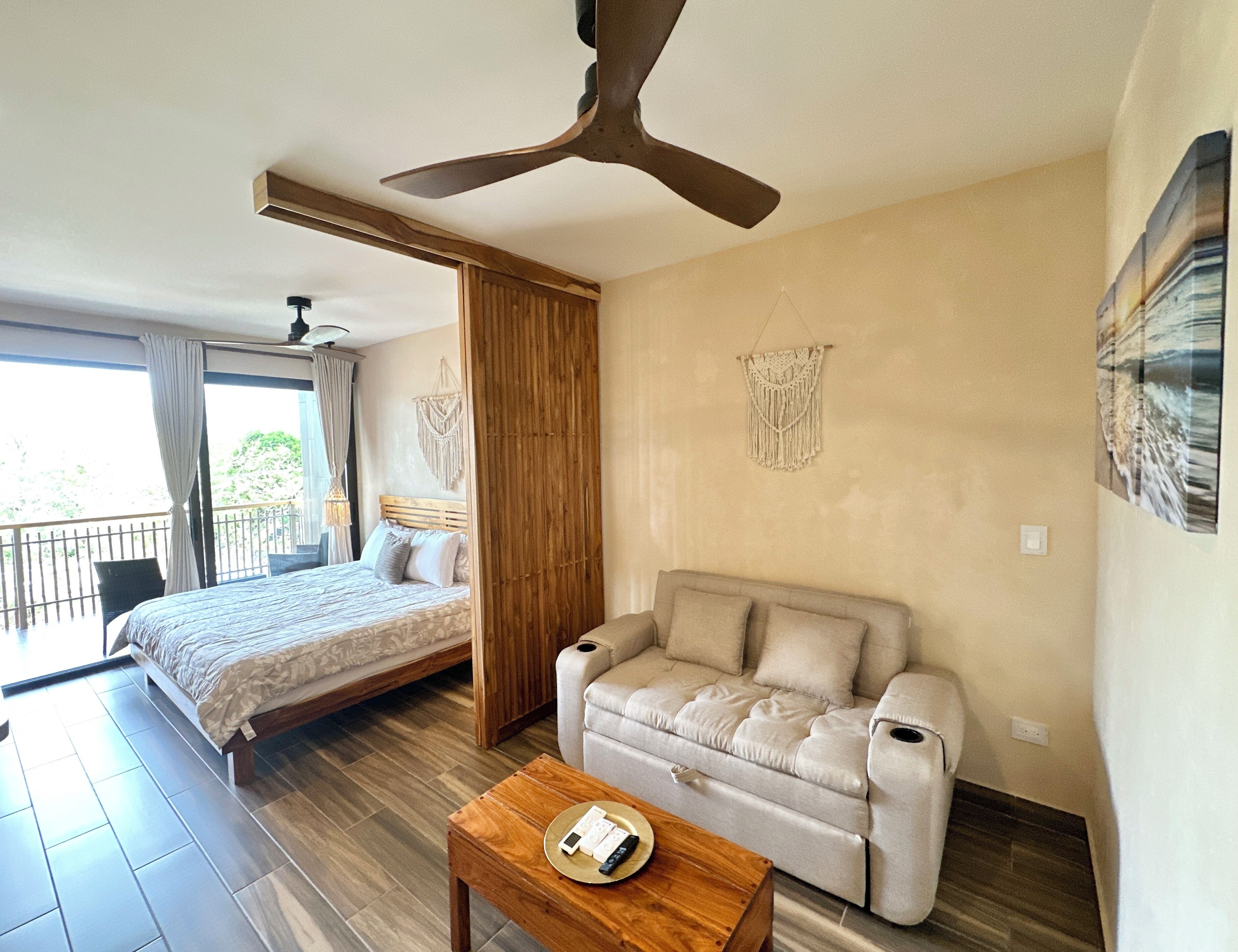 Une chambre d’hôtel moderne équipée d’un lit, d’un canapé, d’une table basse en bois, d’un ventilateur de plafond et d’un balcon avec des rideaux.