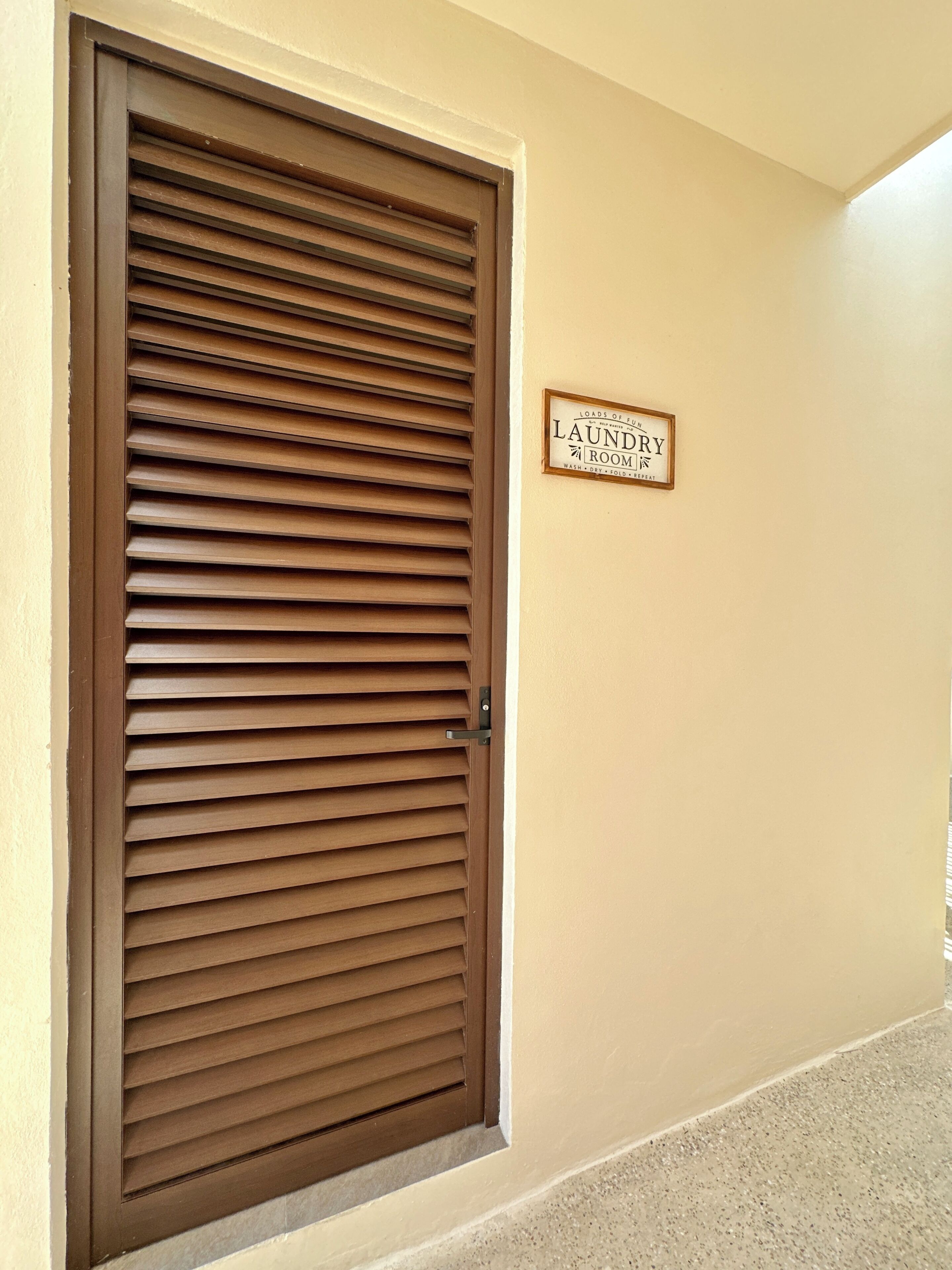 Eine geschlossene Tür mit horizontalen Lamellen, ein Schild darüber mit der Aufschrift 'Laundry Room' und eine beige Wand mit strukturierter Oberfläche.