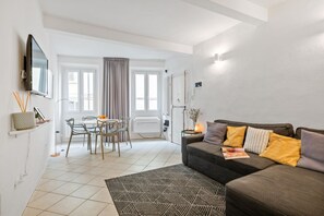 Ein modernes Wohnzimmer mit einer Couch, einem Esstisch mit Stühlen, einem an der Wand befestigten Fernseher und einem großen Fenster mit Vorhängen.