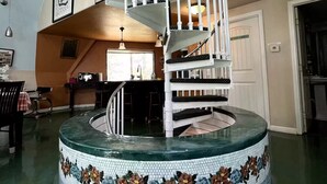 Uma escadaria em espiral com base em azulejos, uma área de jantar com mesa e cadeiras, e uma cozinha ao fundo.