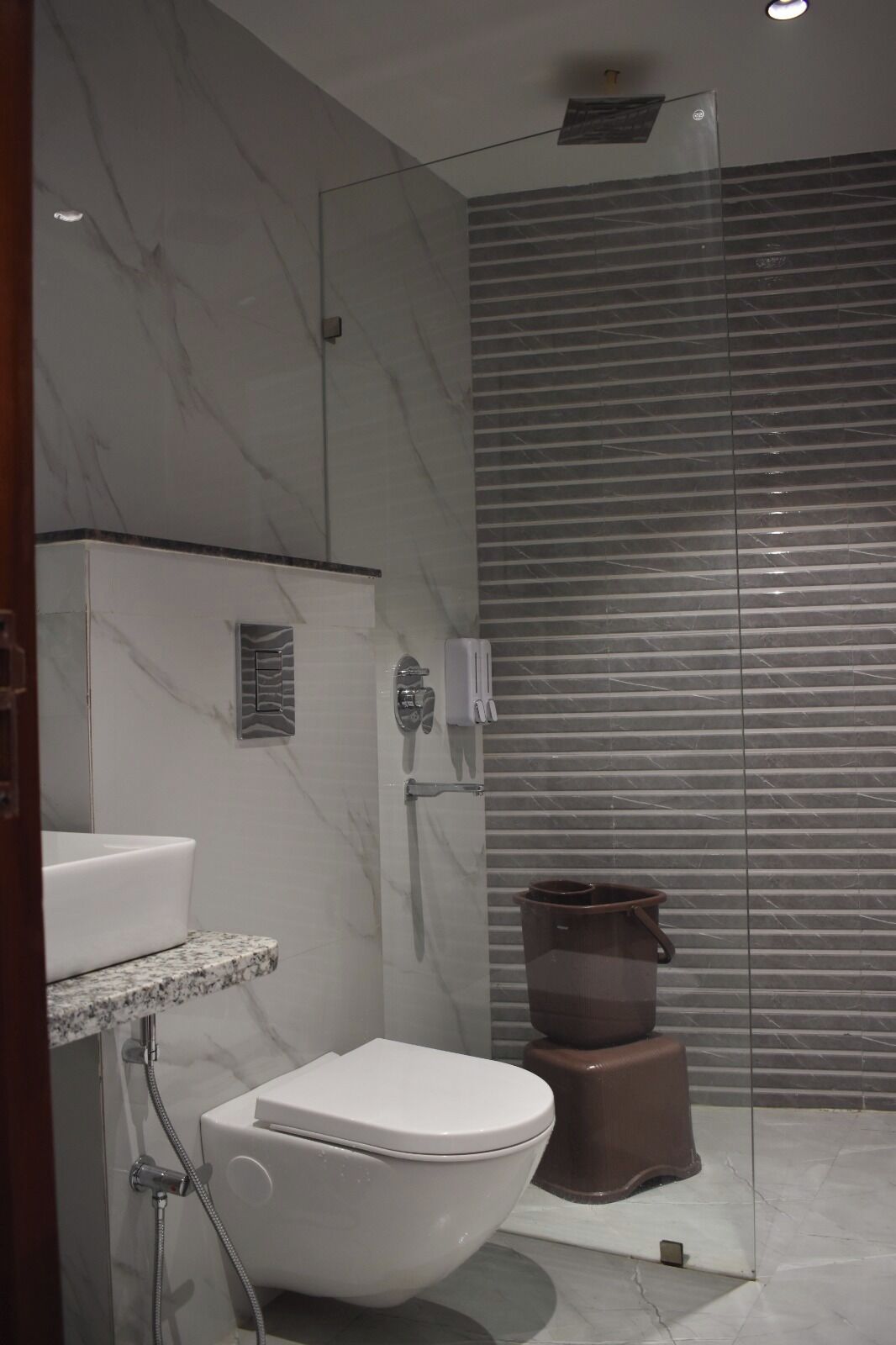 A modern bathroom with a marble wall, a white toilet, a glass shower enclosure, and a trash bin.