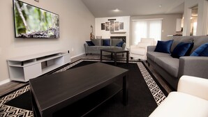 A modern living room with a flat-screen TV, grey sofas with blue cushions, a black coffee table, and a patterned rug. - Full Family Stay Near At&t, Ballpark, 6 Flags & Airport (Grand Prairie)