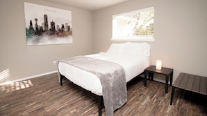 A bedroom with a bed, a bedside table with a lamp, a window with blinds, and a cityscape painting on the wall. - Full Family Stay Near At&t, Ballpark, 6 Flags & Airport (Grand Prairie)