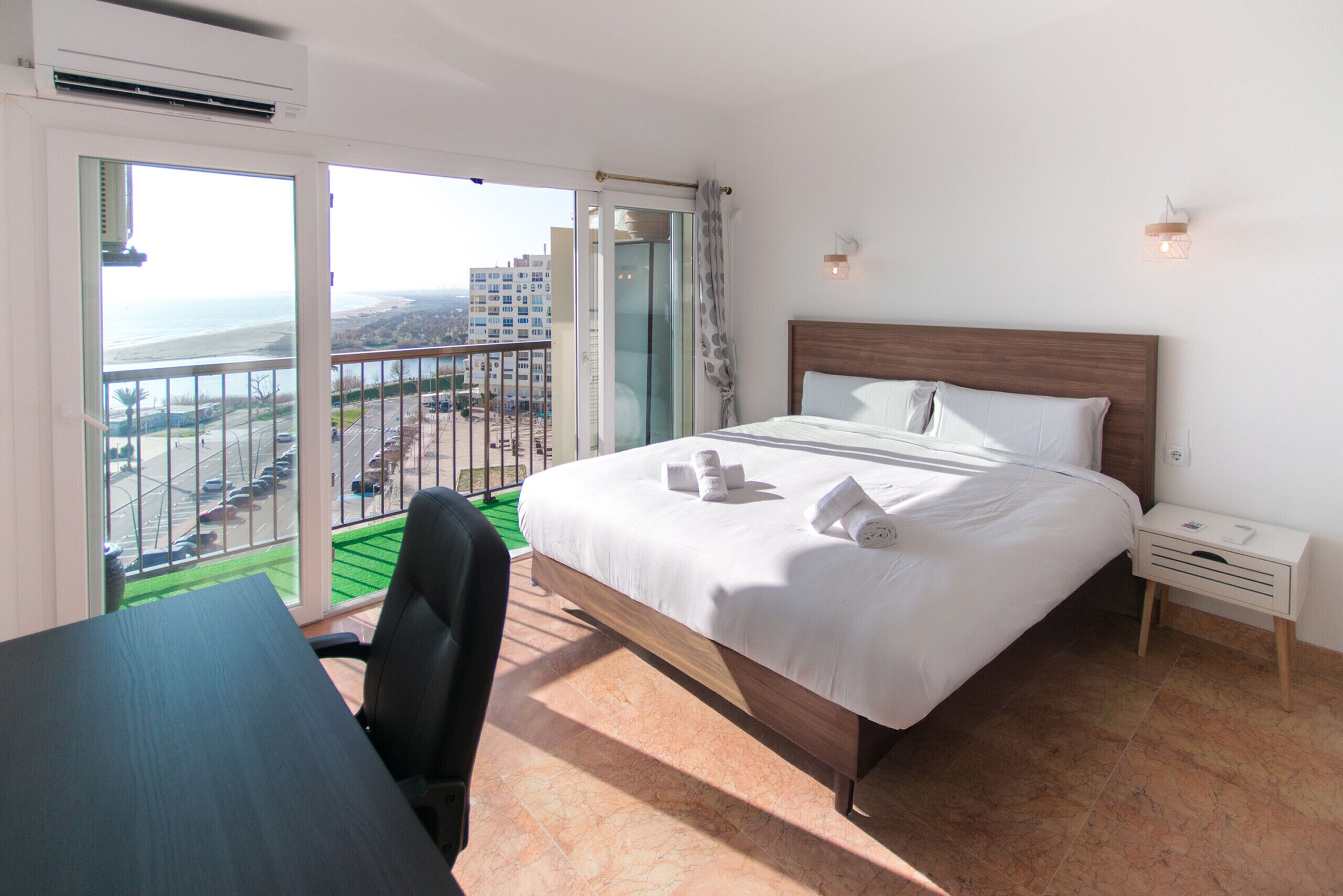 Habitación de hotel con cama, escritorio, silla, mesita de noche y vista al océano desde un balcón.