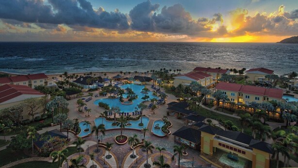 Ein Resort mit einem zentral gelegenen Schwimmbad, mehreren Gästezimmern und Blick auf den Sonnenuntergang über dem Meer.