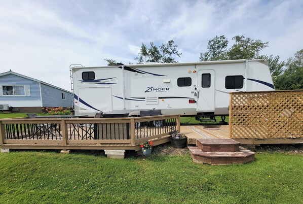 A Zinger Crossroads RV parked in a grassy area with a wooden deck and a wooden fence. - Little Cape RV near harbor and beaches, front bedroom and rear bunk room sleep 6 (Beaubassin East)