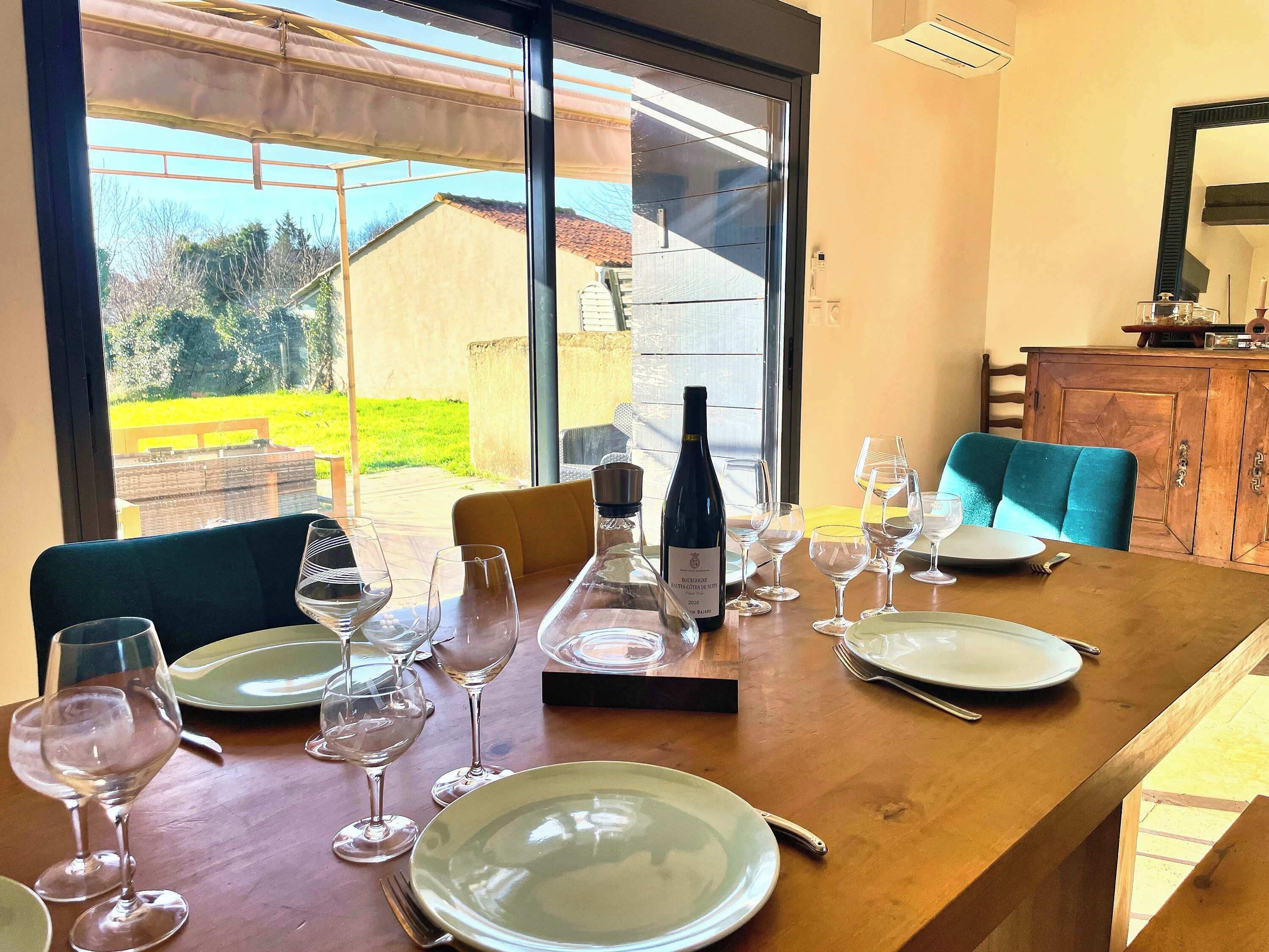 Mesa de comedor dispuesta con copas de vino, platos y una botella, con vista a un jardín y un edificio.
