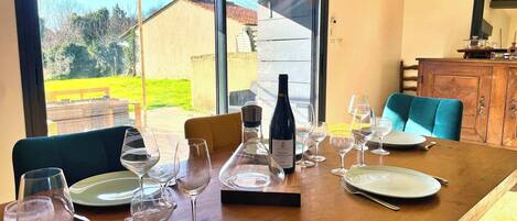 Mesa de comedor dispuesta con copas de vino, platos y una botella, con vista a un jardín y un edificio.