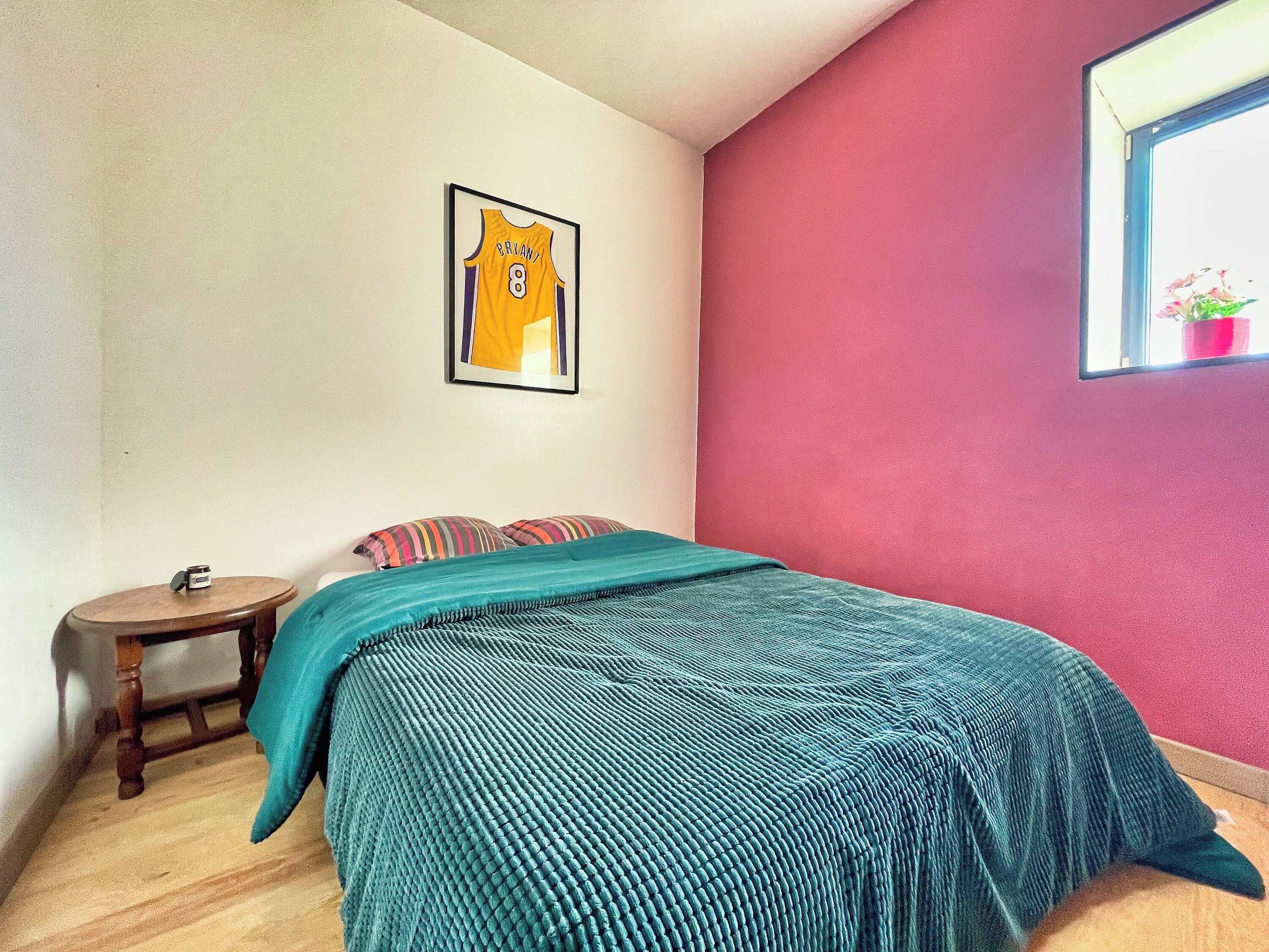 A bedroom with a bed, a framed sports jersey on the wall, a small wooden table, and a window with a plant.