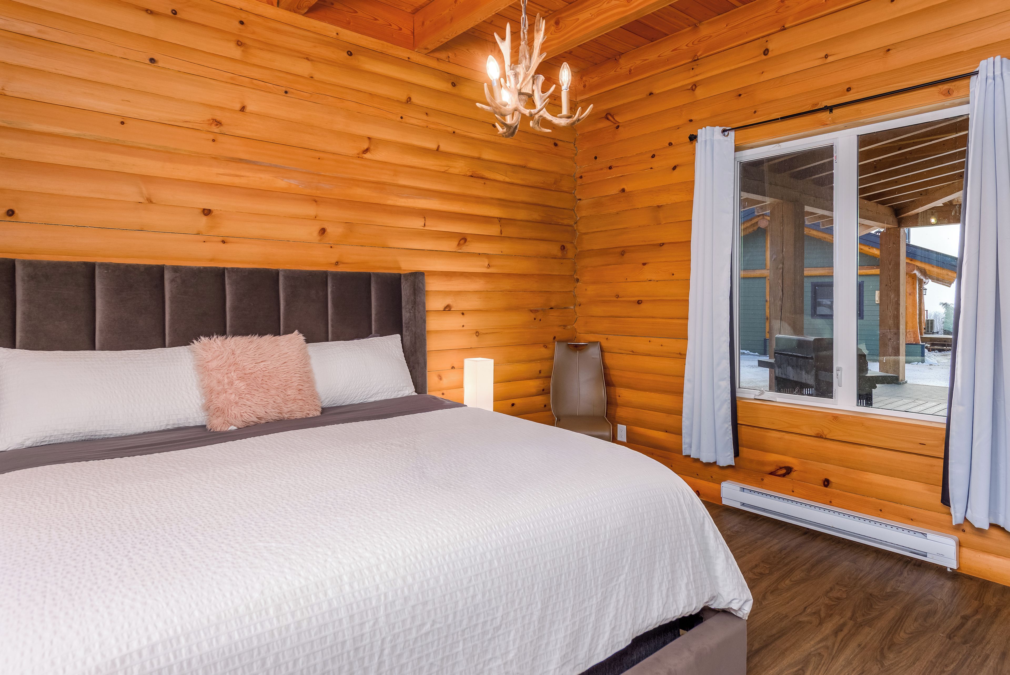 Een slaapkamer in een houten chalet, met een bed, nachtkastje, lamp en uitzicht door een raam.