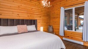 Quarto de cabana de madeira com uma cama, uma mesinha de cabeceira, um abajur e vista pela janela.