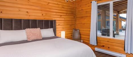 Un dormitorio de cabaña de madera con una cama, una mesita de noche, una lámpara y vistas por una ventana.