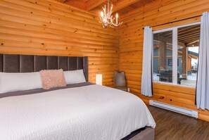 A wooden cabin bedroom with a bed, a nightstand, a lamp, and a view through a window. - Cabin # 2 - Lakeside cabin sleeps 11 with private hot tub (Naramata)