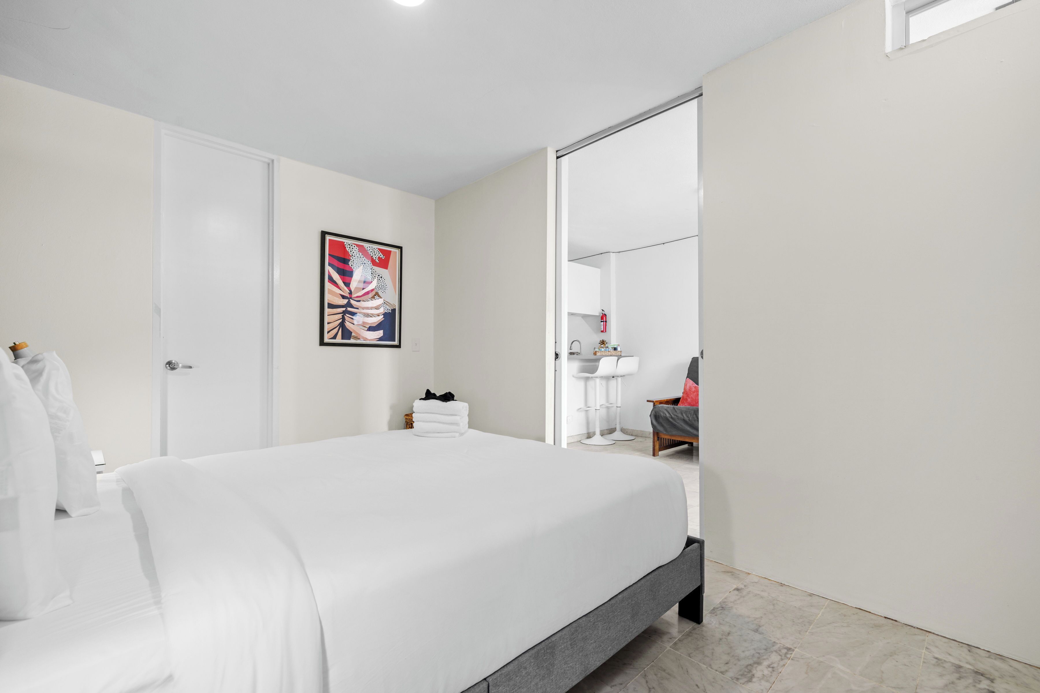 Een hotelkamer met een groot bed, een witte deur, een schilderij aan de muur, een badkamer met een wastafel en spiegel, en een stoel.