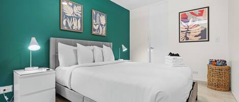 Una cama bien hecha con ropa blanca, un cabecero gris y dos láminas enmarcadas con motivos botánicos en una pared verde azulada.