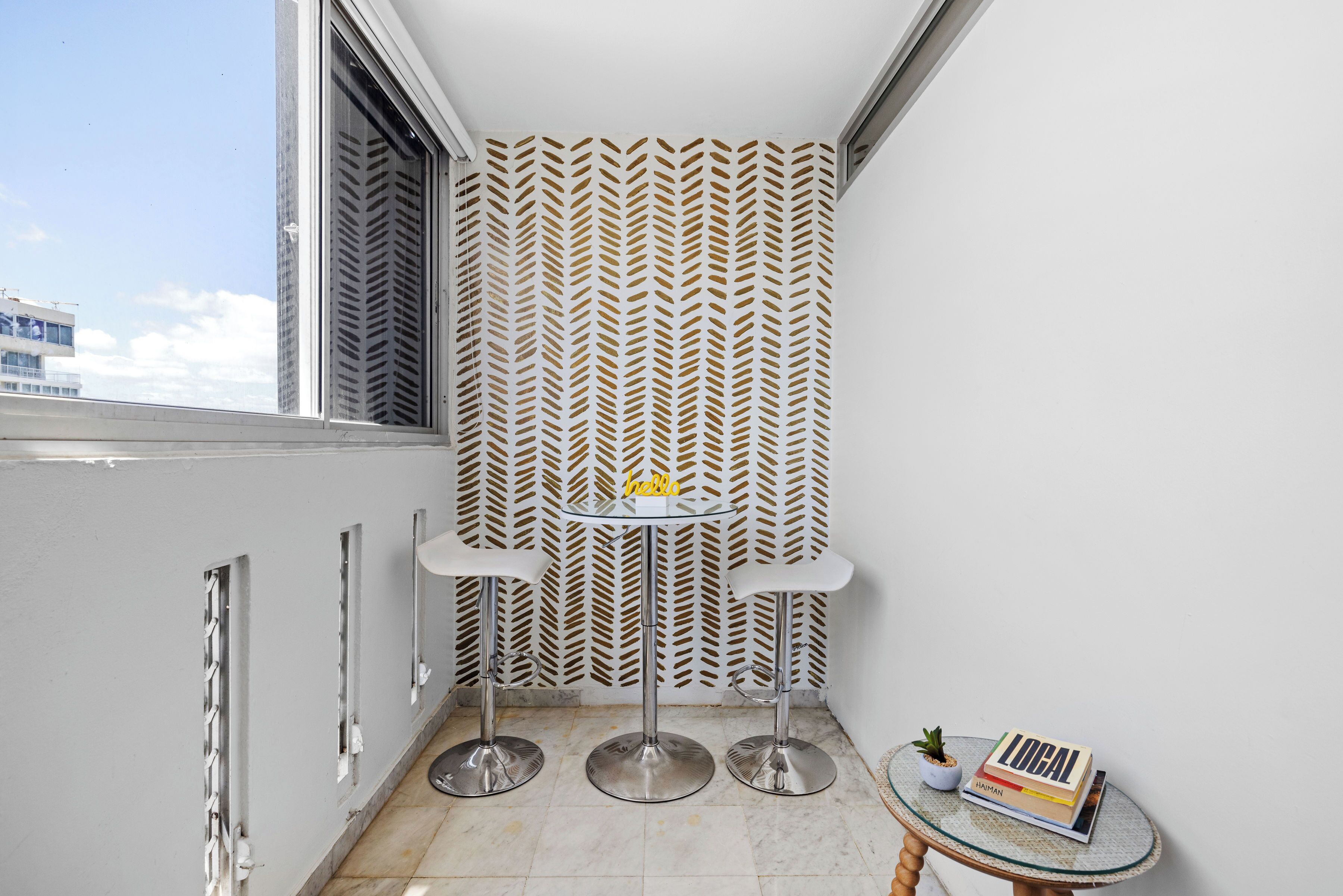 A corner room with a patterned wall, a small round table with two chairs, a potted plant, and a stack of books.