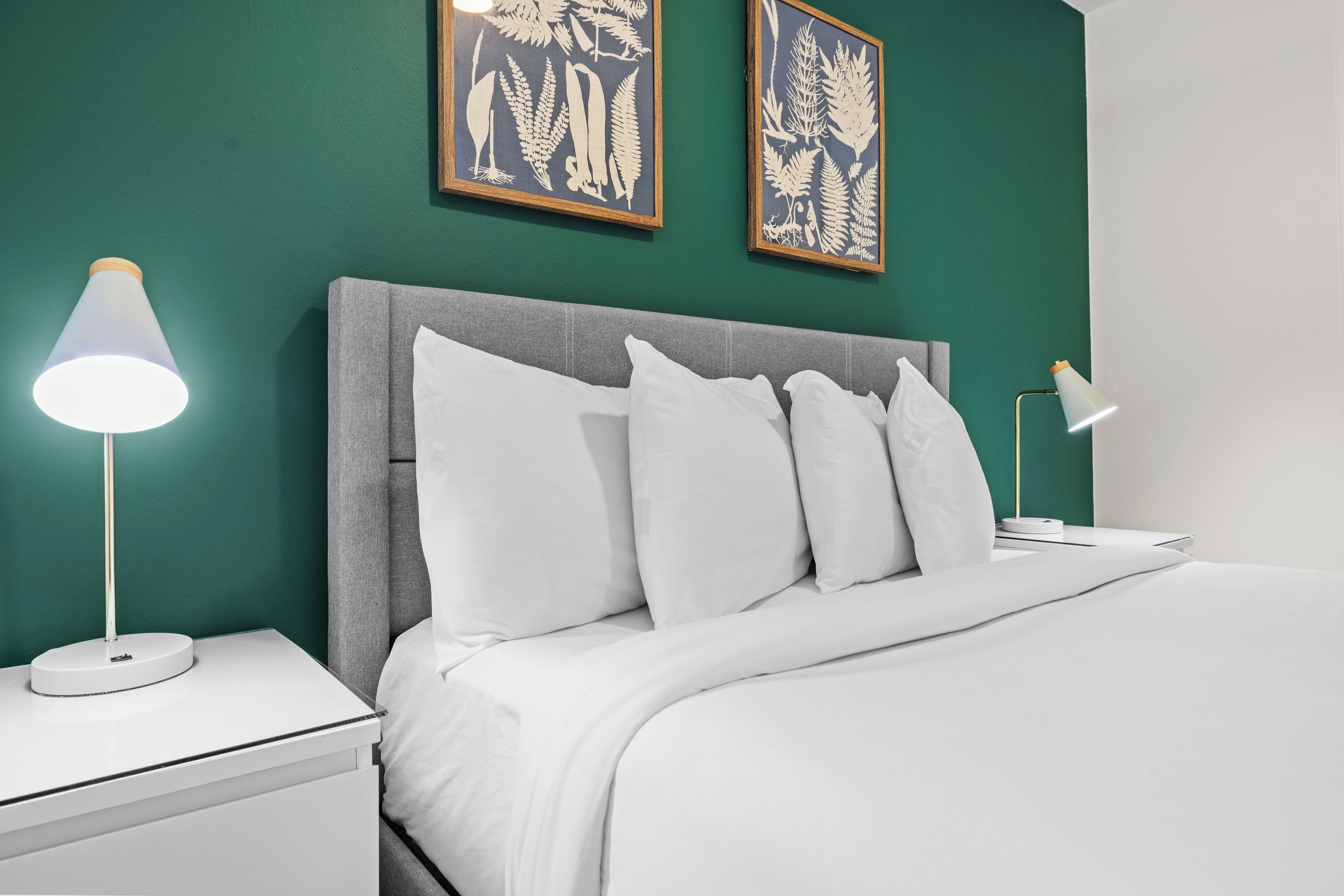 A neatly made bed with white linens, a grey headboard, and two framed botanical prints on the wall.