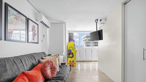 A modern living room with a grey sofa, red and brown cushions, a flat-screen TV mounted on the wall, and a large window offering a view of the cityscape. - Condado del Mar by Otium (San Juan)