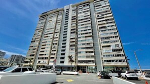 A multi-story building with a modern architectural design, featuring a prominent entrance and a parking area in front. - Condado del Mar by Otium (San Juan)
