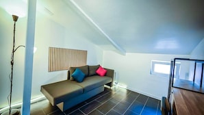 A modern loft apartment with a dark wood floor, a brown sofa with colorful cushions, a tall floor lamp, and a wooden ceiling.