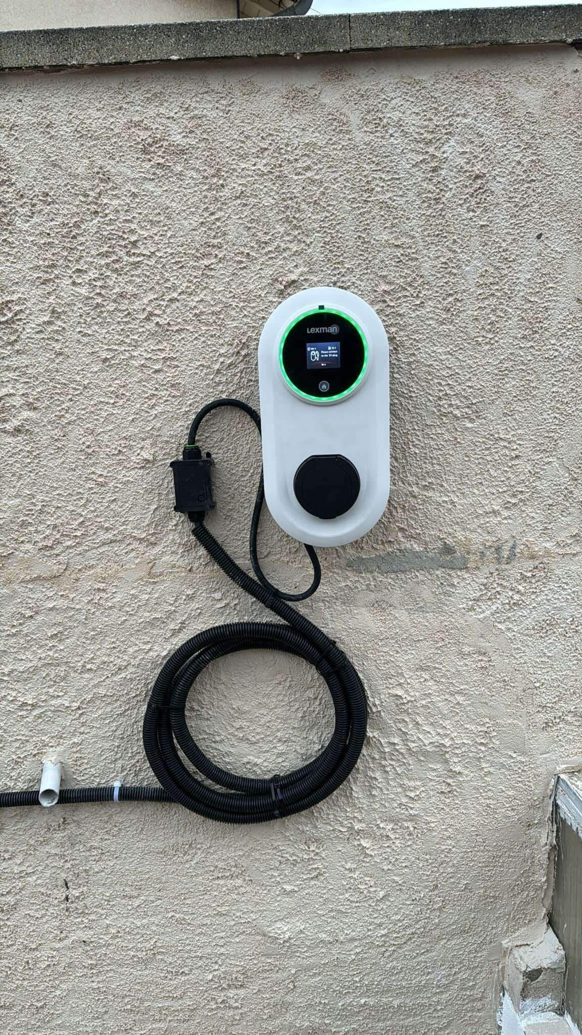 A white Lexman device mounted on a textured wall, connected to a coiled black cable.