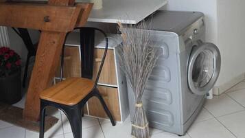 Un área de lavandería con lavadora, escoba, silla y estante con objetos.