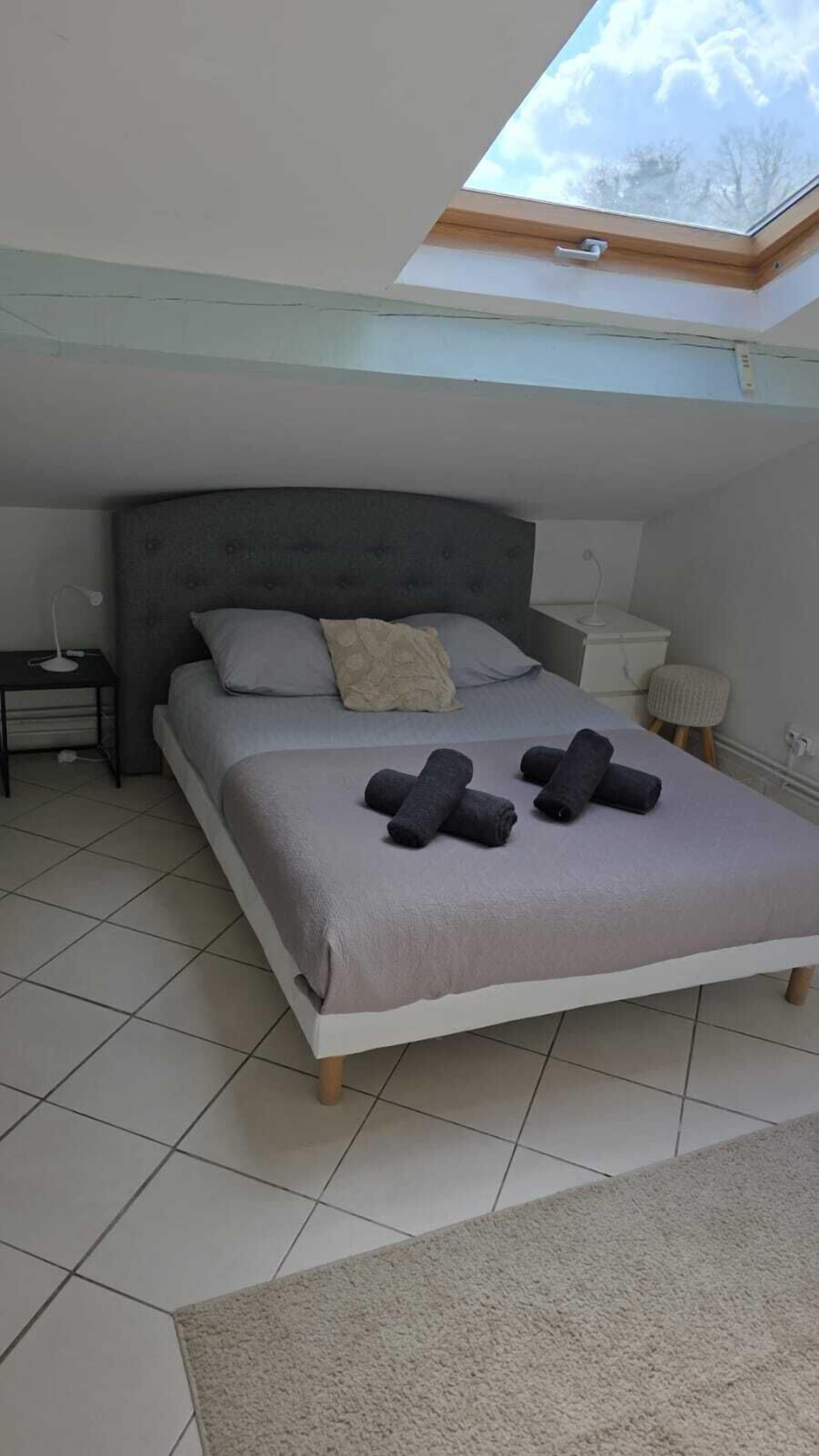 Une chambre avec un puits de lumière, un lit avec des oreillers, une table de chevet, une commode, une lampe et un petit tabouret.