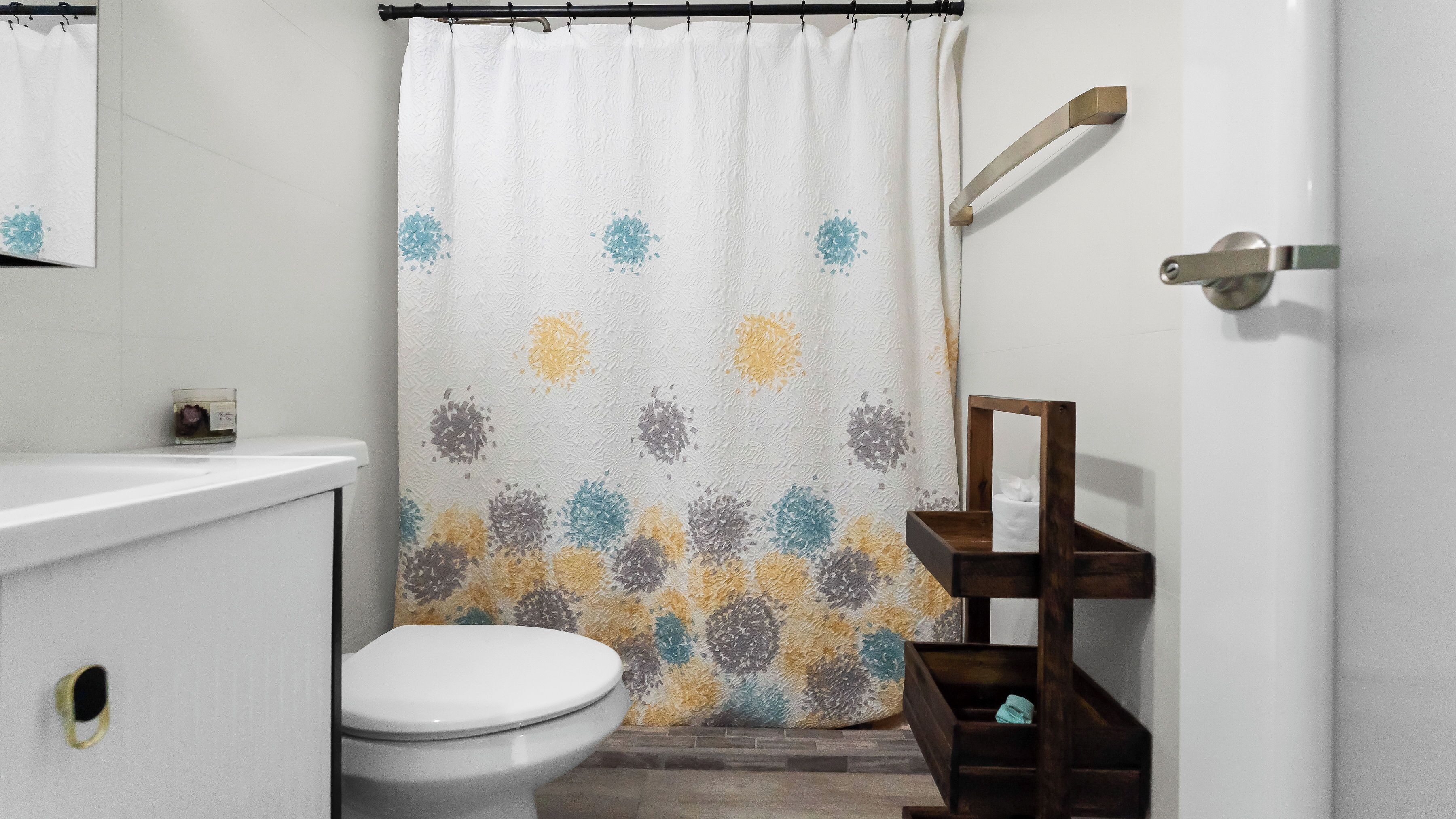A bathroom with a shower curtain featuring a floral pattern, a white toilet, a wooden shelf with a mirror, and a white sink.