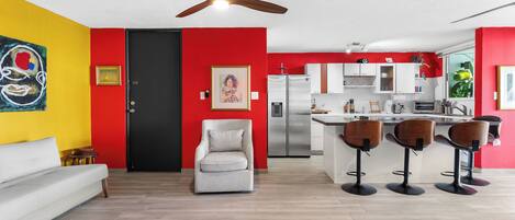 A modern kitchen with a refrigerator, microwave, and sink. There is a dining area with bar stools and a sofa.