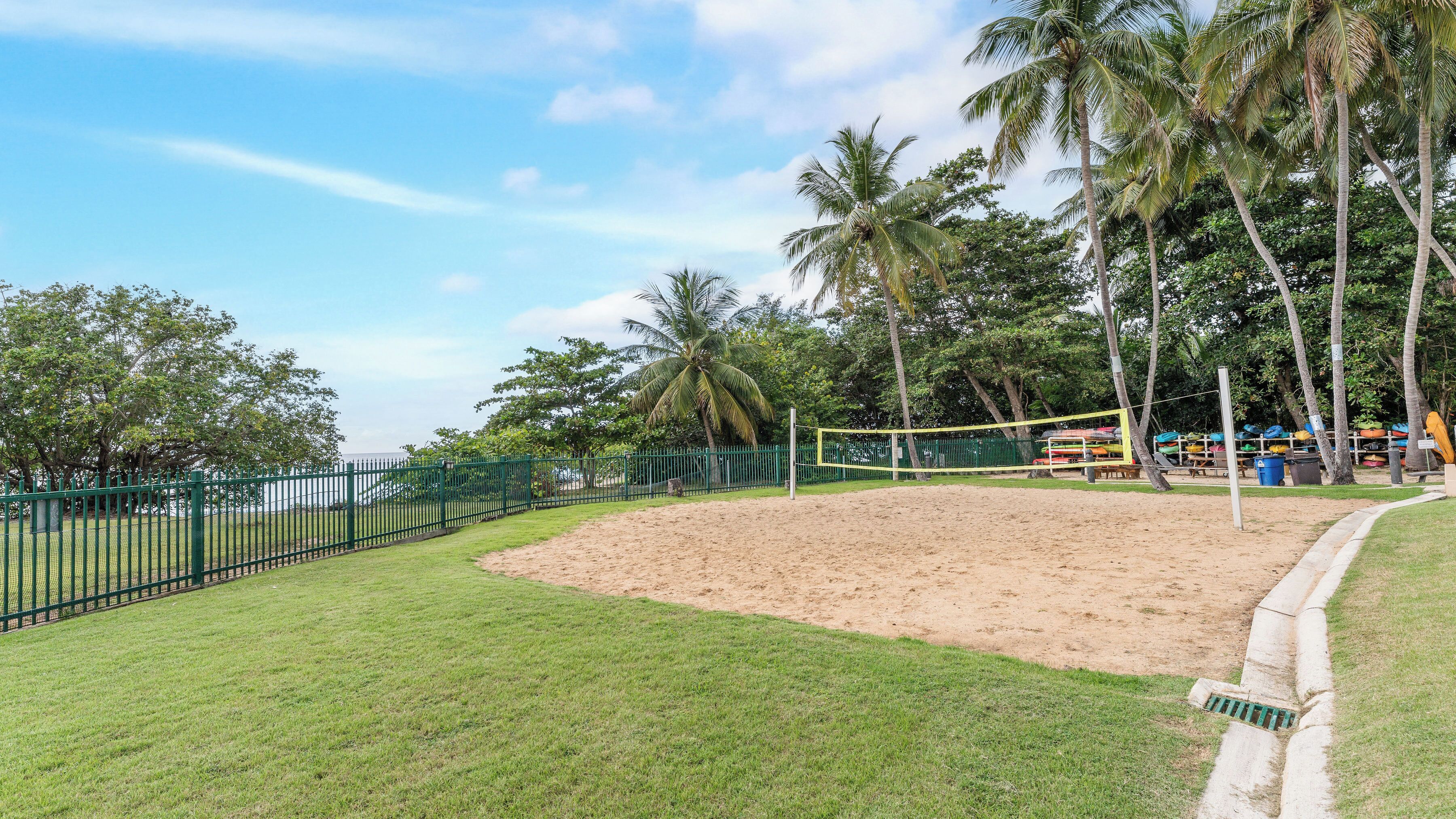 Una cancha de voleibol de arena rodeada por una cerca verde, con palmeras y un área cubierta de césped.
